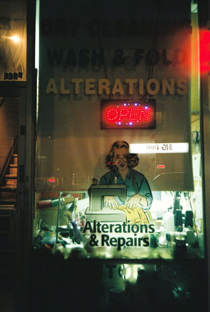 A window view of a Toronto alterations shop, featuring neon signs and workshop tools at night.